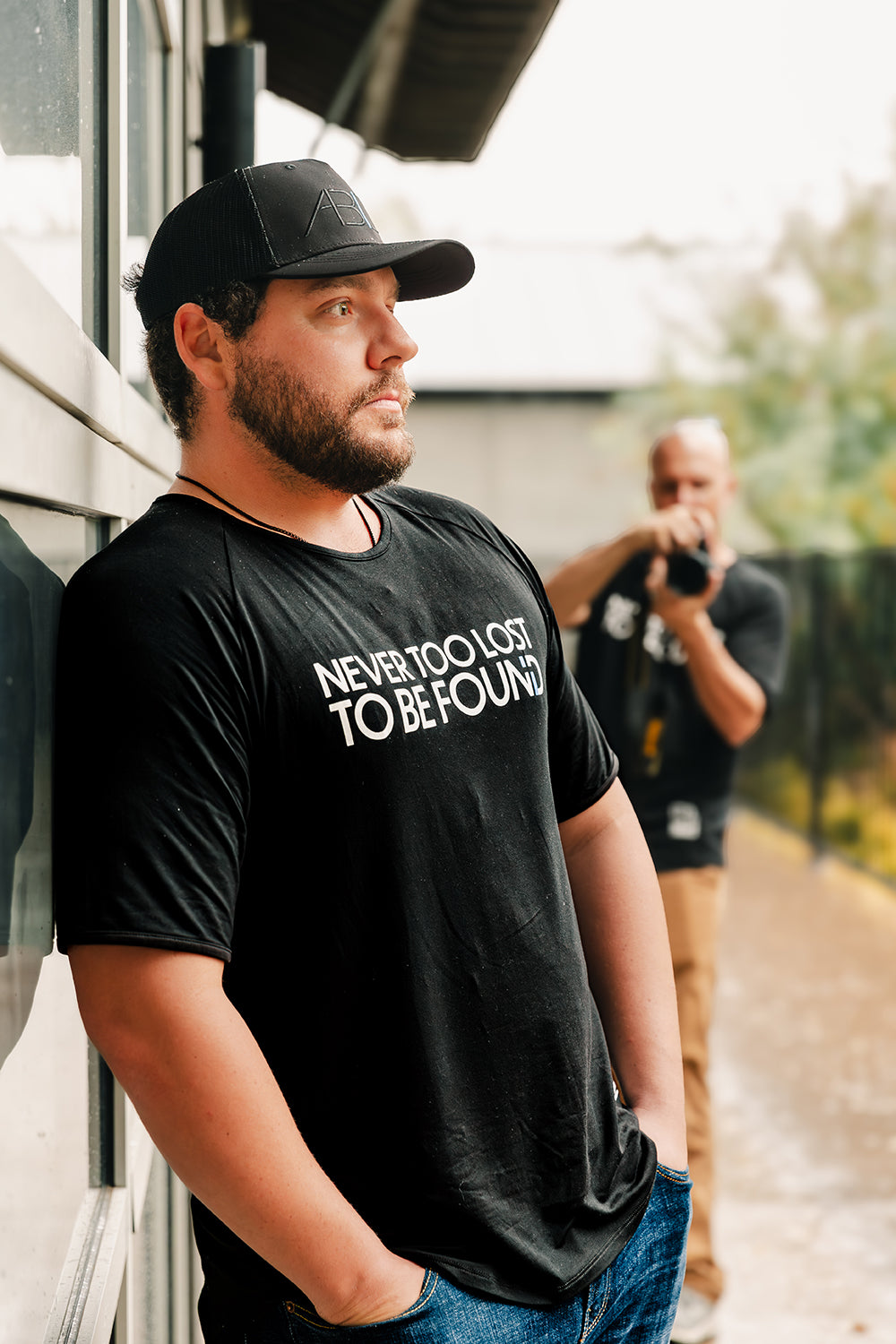 man in "abide" hat and "never too lost" shirt leaning against garage door, looking into distance, photographer blurred in background.