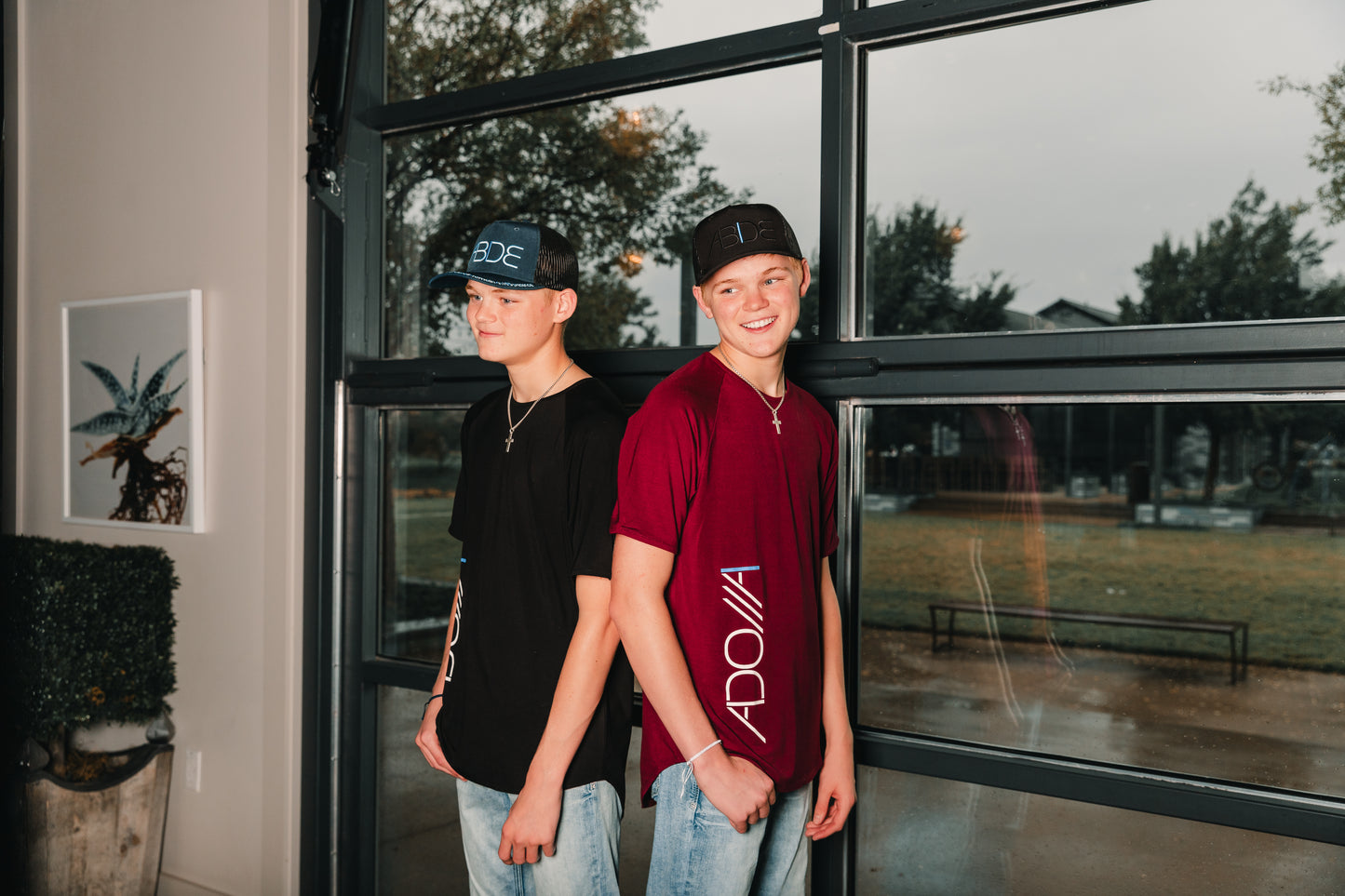 two teen boys wearing now found christian apparel "adonai" bamboo tees and "abide" trucker hats, one in ocean blue and black, the other in black and wine red.