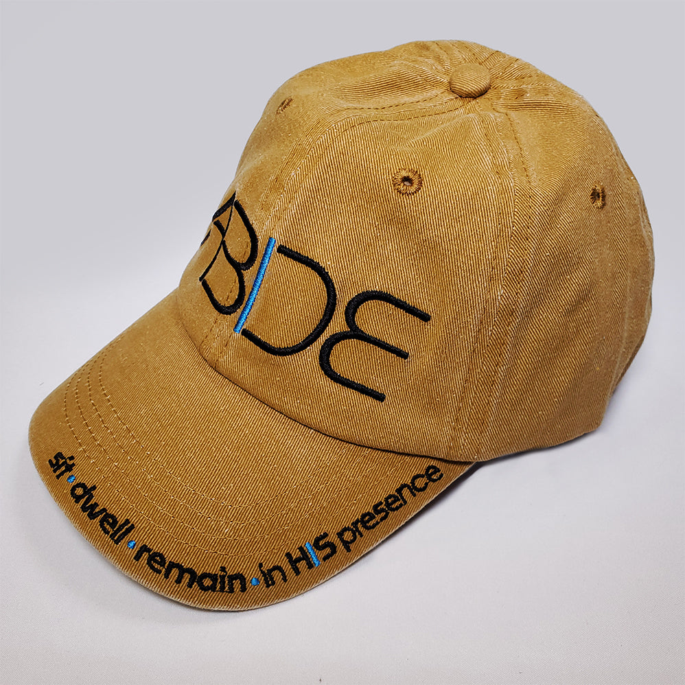 A mustard yellow dad hat with "Abide" embroidered on the front and Sit - dwell - remain in His presence on the bill from a front left perspective