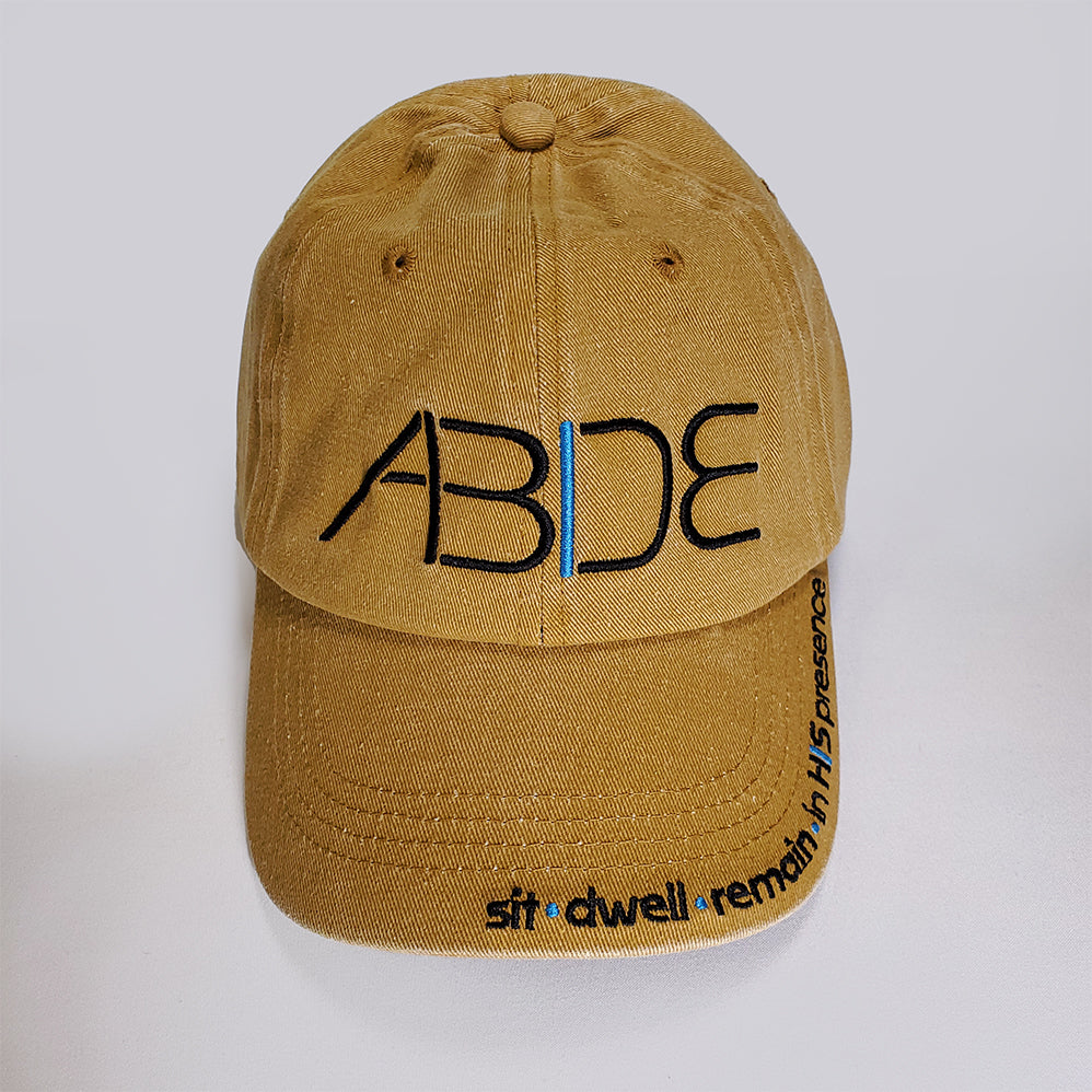 A mustard yellow dad hat with the word "Abide" embroidered on the front and sit - dwell - remain in His presence on the bill